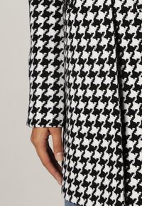 Houndstooth-mönstrad kappa med en svart och vit färgschema, tillverkad av texturerad tyg. Har en figursydd silhuett och långa ärmar.