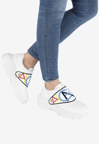 Des baskets blanches à semelle épaisse présentent une bride colorée affichant "AG" en bleu, rouge et jaune sur un fond noir.