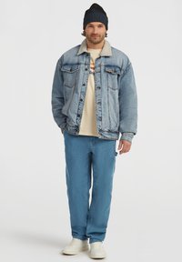 Veste en denim bleu clair avec un col en shearling blanc, assortie de pantalons bleus clairs et de baskets blanches, le tout porté par un homme portant un bonnet noir.