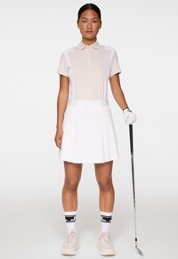 Kobieta w beżowo-białym stroju golfowym trzymająca kij golfowy, stojąca na tle jednolitym.