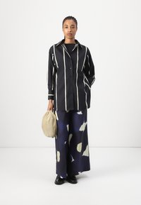 Black and white striped shirt with a button placket, paired with wide-leg navy pants featuring abstract light-colored patterns, holding a beige bag.