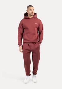 Vinröd hoodie med framficka och dragsko, matchas med likadana joggers. Vita sneakers fulländar outfiten. Mjukt, strukturerat material.