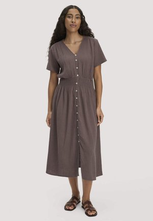 Frau mit langen, lockigen Haaren trägt ein braunes Midikleid mit kurzen Ärmeln und durchgehender Knopfleiste sowie braune Sandalen, steht vor einem neutralen Hintergrund.