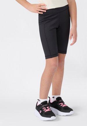Enfant portant un short de sport noir jusqu'aux genoux, des chaussettes blanches Champion et des baskets noires avec des lacets roses et des semelles blanches, se tenant devant un fond blanc.