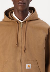 Haut à capuche zippé marron en tissu mélangé coton, avec un cordon de serrage, une grande poche kangourou à l'avant et un petit logo cousu sur la poitrine.