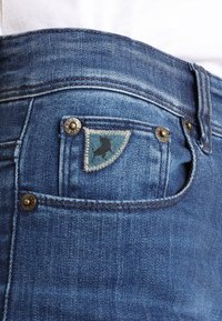 Modré džíny vyrobené z denimu s charakteristickou trojuholníkovou náprsní kapsou so šitím. Kovové prvky pri kapsách a opasku.