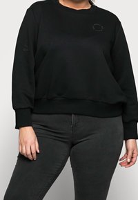 Zwarte sweatshirt met een ronde hals, ribgebreide manchetten en een klein logo-detail. Gepaard met donkergrijze broek voor een gecoördineerde uitstraling.