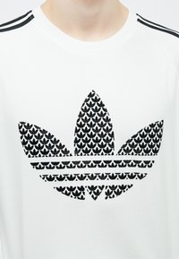 Fehér póló egy feltűnő fekete Adidas logóval, amely levélmintát és mindkét vállon három csíkot ábrázol. Puha anyag, környakú kivágás.