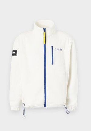 Bela flis jakna z modrim zadrgo, žepom na prsih, nastavljivimi vrvicami na robu, logotipom Red Bull Racing na prsih in črno nalepko na rokavu.