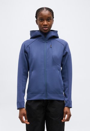 Γυναίκα με πλεξούδες, φοράει μπλε ζακέτα με κουκούλα και φερμουάρ, καθώς και μαύρο παντελόνι, στέκεται μπροστά σε απλό ανοιχτόχρωμο φόντο.