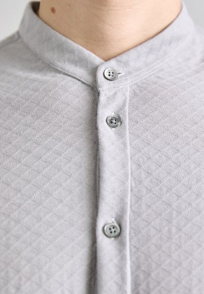 Ljusgrå krag skjorta med en texturerad diamantmönster, med tre grå knappar vid halsen och en slät tygtextur.