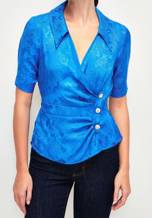 Woman wearing a short-sleeve blue floral wrap blouse with a wide collar and three gold buttons, paired with dark denim jeans.