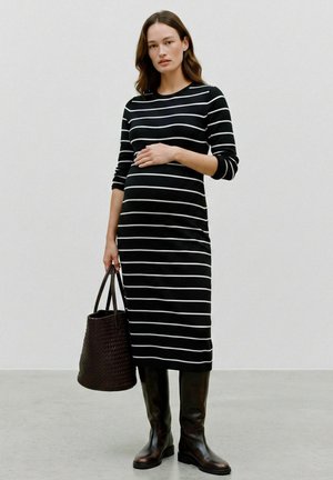 Femme enceinte portant une longue robe rayée noire et blanche et des bottes noires, tient un sac à main tissé foncé d'une main sur son ventre.