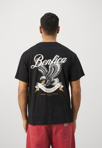 T-shirt en coton noir avec un grand graphique d'aigle et le texte "Benfica" en blanc et or, avec un design de ruban courbé en bas.