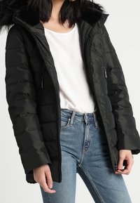 Svart puffjacka med huva och fuskpälsdetaljer, med dragkedjefickor och en quiltad design, bärs över en vit skjorta och blå jeans.