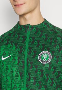 Chaqueta de chándal con cremallera en verde, con un patrón geométrico de triángulos, logo de Nike y un distintivo circular de la Federación Nigeriana de Fútbol.