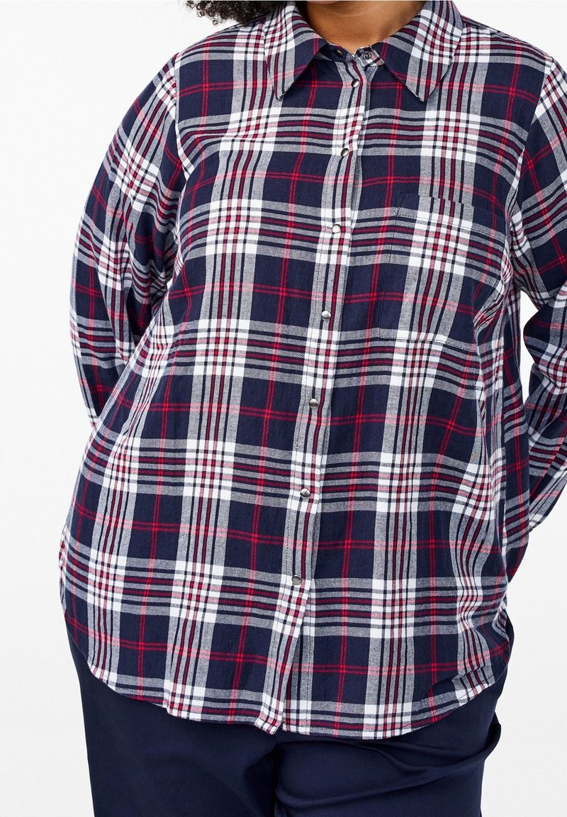 Personne portant une chemise à carreaux bleu marine, blanc et rouge, à manches longues, boutonnée, avec un pantalon foncé, mains derrière le dos, cadrée au niveau du cou et des cuisses.