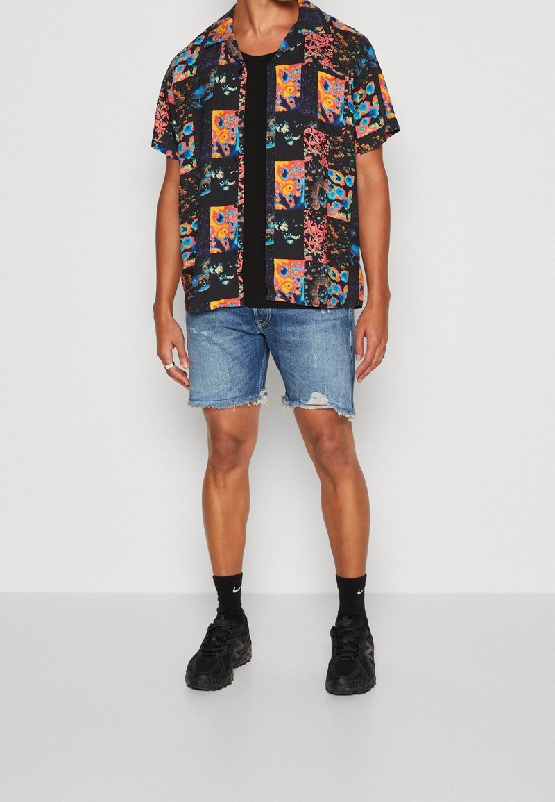 Camicia a maniche corte con motivi floreali e geometrici colorati su sfondo nero, abbinata a pantaloncini di jeans strappati e scarpe da ginnastica nere.