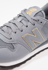 Baskets New Balance 500 gris avec des lacets gris, des matériaux en mesh et en daim, un logo doré sur la languette et un grand « N » cousu sur le côté.