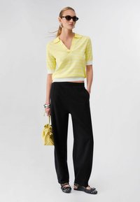 Femme portant un pantalon noir, un haut à manches courtes rayé jaune, des ballerines noires cloutées, des lunettes de soleil noires, des boucles d'oreilles argentées, et un sac à main jaune.