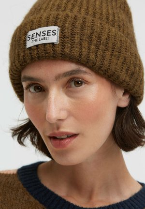 Woman with short dark hair wearing a brown ribbed knit beanie labeled "SENSES THE LABEL" and a brown sweater with a navy collar.