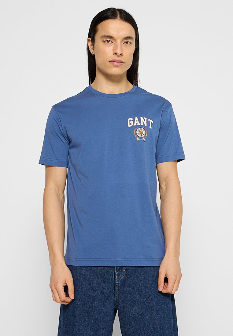 Blaues Baumwoll-T-Shirt mit rundem Halsausschnitt und kurzen Ärmeln. Verfügt über ein kreisförmiges Logo mit dem Wort "GANT" in crèmefarbener Schrift.