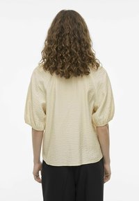 Top de tela arrugada color beige claro con mangas abullonadas, un ajuste relajado y dobladillo redondeado. La vista trasera no muestra cierres ni adornos.