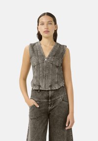 Top de denims sin mangas en negro y gris, con un escote en forma de V profundo, cierre de cremallera y tejido texturizado. Combinado con pantalones holgados a juego.