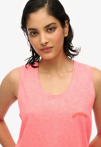 Różowy bezrękawnik wykonany z lekkiego materiału, z okrągłym dekoltem i logo "Superdry" w kontrastowym haftowanym wzorze.