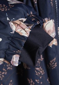 Veste bleu marine avec motif floral ; dotée de poignets côtelés et d'un tissu doux et texturé. Gros plan montrant le détail de la manche froncée.