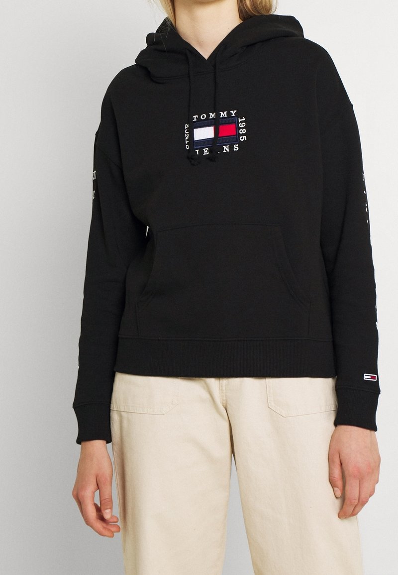 Personne portant un sweat à capuche noir Tommy Jeans avec logo sur la poitrine et la manche, associé à un pantalon beige clair, debout devant un fond blanc uni.