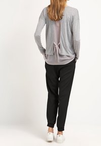 Nainen nähtynä takaa, päällään harmaa pitkähihaine paita avonaisella selällä, joka on solmittu vyötärölle, mustat housut ja valkoiset lenkkarit.