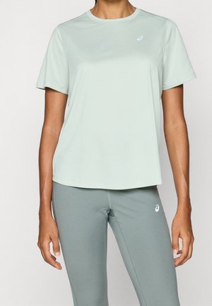 Frau trägt hellgrünes kurzärmeliges Sportshirt und passende Leggings, zeigt Oberkörper und Arme vor einfarbigem Hintergrund.