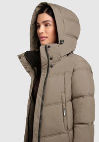 Beige pufferjack met een gequilte ontwerp, een grote capuchon, een voorrits en drukknoopjes. De binnenkant heeft een donkere voering.