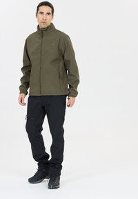 Oliven grøn softshell jakke med høj krave, frontlynlås og sidelommer, parret med sorte tapered bukser og sorte vandresko.