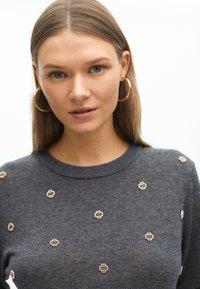 Grauer Pullover mit Rundhalsausschnitt, verziert mit kleinen metallischen Details in Goldfarbe auf der Vorderseite. Glatte Textur und figurbetonende Silhouette.