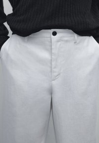 Weiße Leinenhosen mit lockerem Schnitt, ausgestattet mit einem Knopfverschluss und Seitentaschen, kombiniert mit einem strukturierten schwarzen Pullover.