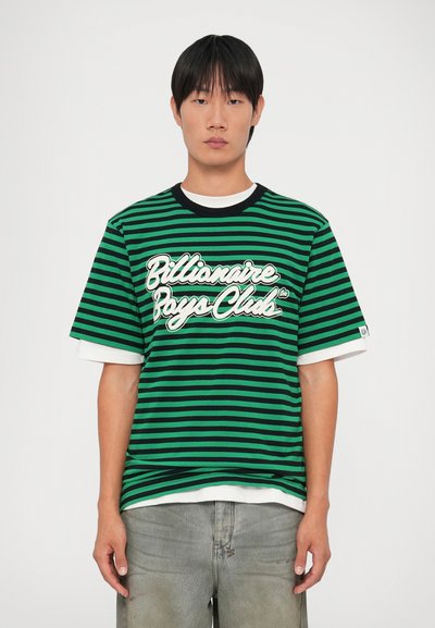 Zelena i crna prugasta majica s okruglim izrezom, na kojoj se nalazi logo "Billionaire Boys Club" u bijeloj boji, složena preko bijele košulje.