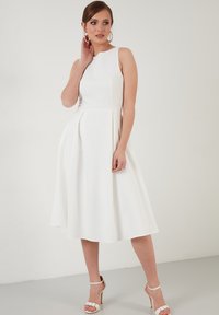Rochie albă fără mâneci, cu un corp bine structurat și fustă evazată. Material neted cu pliseuri; modelul poartă sandale cu barete și cercei cerc.
