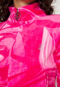 Rosa sammetsjacka med dragkedja och batikmönster, hög krage och strass-logotyp. Har sidofickor med dragkedja och en slät textur.