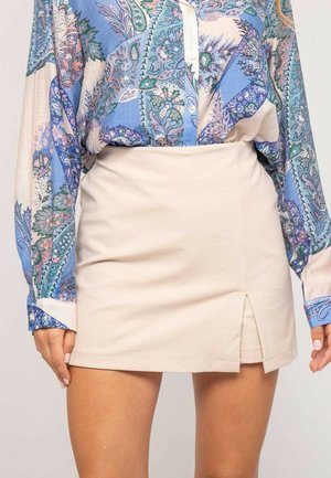Blouse à manches longues bleu à motif cachemire rentrée dans une mini-jupe beige avec une fente à l'avant, vue de la mi-torse jusqu'à la mi-cuisse.