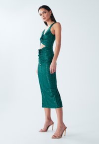 Žalia suknelė su blizgančiomis sequinomis, aptempto silueto, su išpjovomis, reguliuojamomis raištelėmis ir vidutiniškai žemai iki blauzdų. Lygus tekstūra su blizgiu paviršiumi.