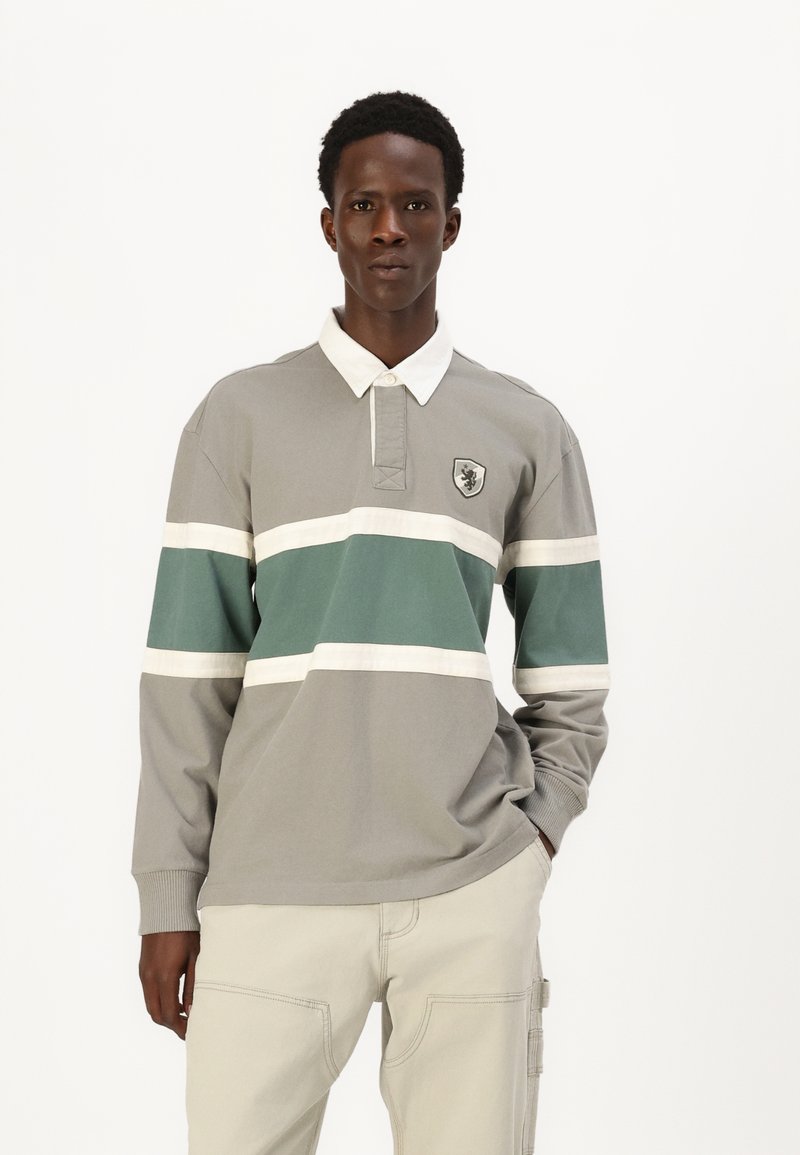 Langærmet polo shirt i grå med grønne og hvide vandrette striber. Har en hvid krave og et løveemblem på brystet. Lavet af bomuldsmateriale.