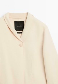 Helle beige Jacke aus weichem Stoff mit Schalkragen, Ein-Knopf-Verschluss und minimalen Nähdetails. Markenetikett ist innen sichtbar.