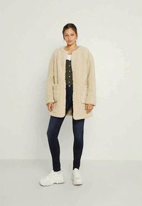 Beige, huňatý, oversized kabát vyrobený z umělé kožešiny, s dlouhými rukávy a dvěma předními kapsami. Kombinovaný se zeleným topem a tmavými džínami.