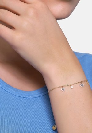 Delikatna złota bransoletka z łańcuszkiem, posiadająca trzy małe zestawy pereł oraz jeden przezroczysty kamyczek, noszona na nadgarstku na tle niebieskiej koszuli.