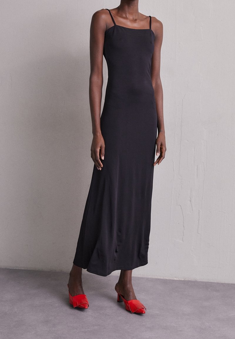 Personne grande portant une robe maxi noire sans manches avec de fines bretelles et des mules à talons rouges avec des détails à franges, debout sur un sol gris.