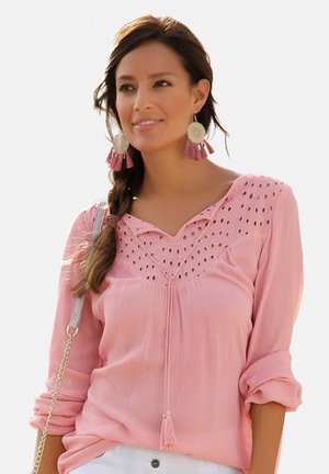 Helles rosa langärmliges Bluse mit Ösenverzierungen, einem V-Ausschnitt und einer gebundenen Quaste vorne. Das Model trägt große runde Ohrringe.
