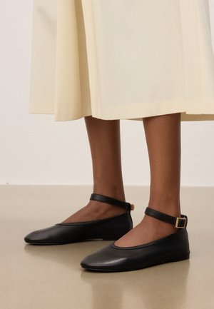 Vue en longueur des jambes d'une personne portant une jupe midi beige et des ballerines en cuir noir avec des brides à la cheville et des boucles dorées.