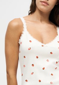 Femme portant un haut blanc sans manches avec bordure en dentelle et petits motifs rouges de fraises sur un fond clair uni.